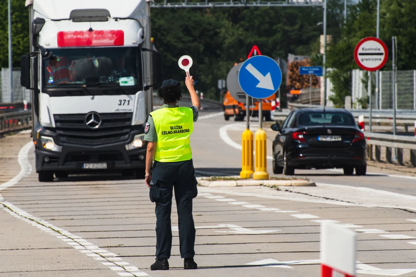 Kontrole na granicach z Niemcami i Litwą. Utrudnienia dla kierowców Funkcjonariusz Służby Celno-Skarbowej kieruje ruchem na przejściu granicznym, zatrzymując ciężarówkę, w tle widoczne znaki drogowe oraz samochody osobowe.