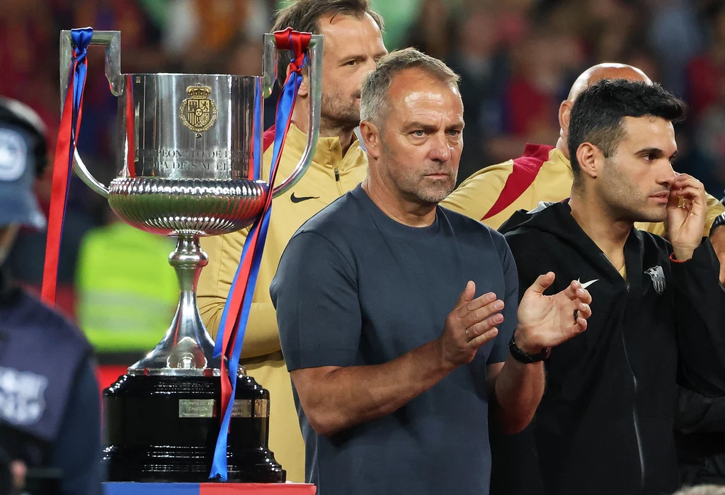 Mężczyzna w ciemnej koszulce stoi obok dużego pucharu sportowego ozdobionego kolorowymi wstążkami, w tle widoczne inne osoby i rozmyty tłum.