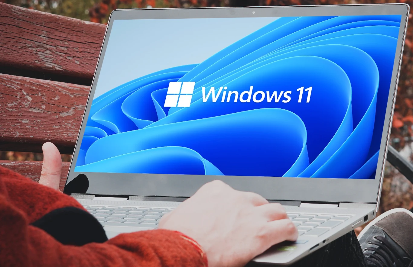 Windows 11 z funkcją z czasów Windows Vista. Obejmuje tapety. Otwartego laptopa z wyświetlonym na ekranie logo Windows 11 oraz charakterystycznym niebieskim tłem, użytkownik trzyma dłoń na klawiaturze, całość na tle drewnianej ławki i rozmytego pleneru.