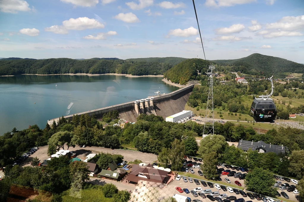 Zapora wodna otoczona zielenią, z dużym sztucznym zbiornikiem wodnym oraz kolejką linową przebiegającą nad kompleksem turystycznym z samochodami i budynkami na pierwszym planie; w tle widać wzgórza i lasy.