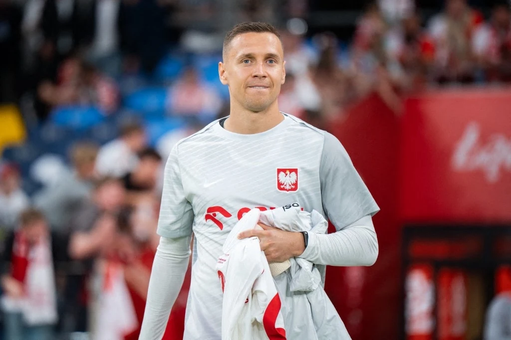 Sportowiec w jasnej koszulce z godłem Polski trzyma w ręku koszulkę, za nim widoczni kibice na trybunach oraz czerwony baner z boku, dominują barwy biało-czerwone.