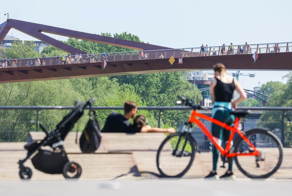 Ze względu na zmianę klimatu wilgotność powietrza może nam częściej dokuczać latem Zacienione miejskie otoczenie z nowoczesnym mostem w tle, na którym spaceruje wiele osób, na pierwszym planie kobieta obok jaskrawego roweru, w pobliżu ławki siedzi para z wózkiem dziecięcym, całość utrzymana w letniej atmosferze.