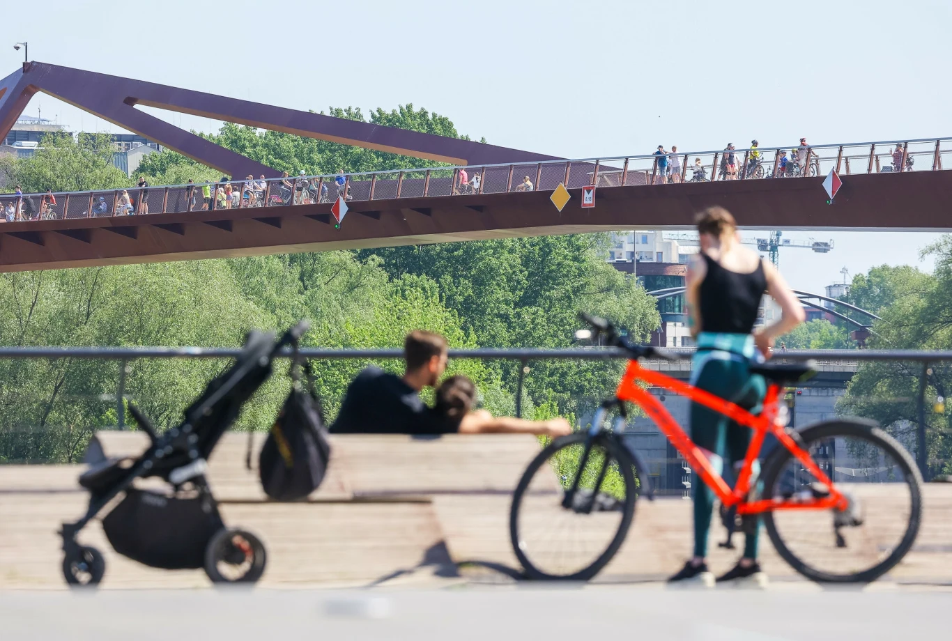 Zacienione miejskie otoczenie z nowoczesnym mostem w tle, na którym spaceruje wiele osób, na pierwszym planie kobieta obok jaskrawego roweru, w pobliżu ławki siedzi para z wózkiem dziecięcym, całość utrzymana w letniej atmosferze.