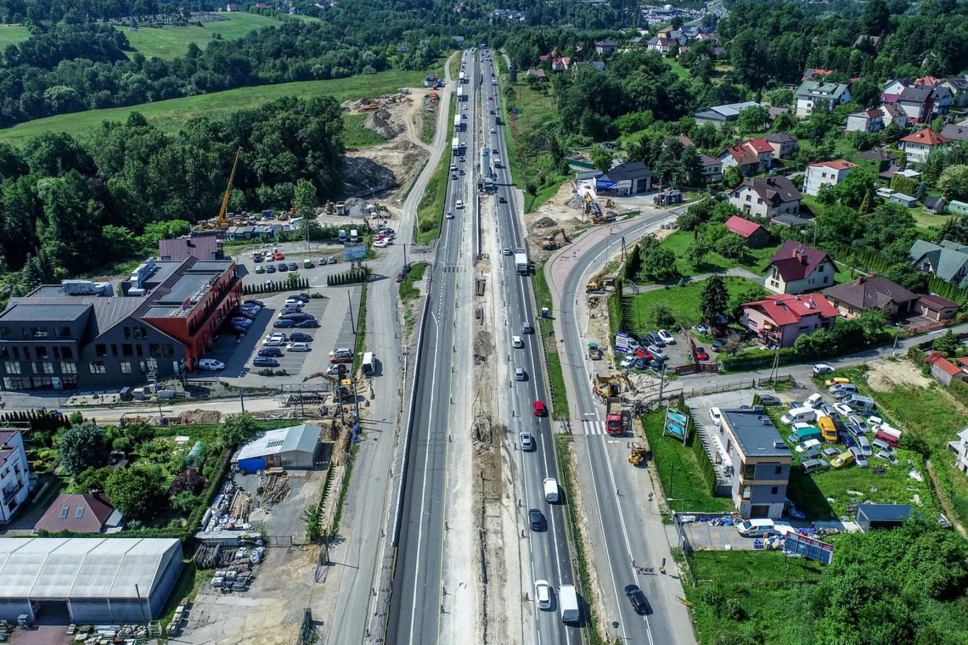 W szczycie wakacyjnych wyjazdów ruszają duże zmiany w organizacji ruchu na jednej z najważniejszych tras w Małopolsce Droga w trakcie rozbudowy, otoczona przez samochody poruszające się po obu stronach, widoczne maszyny budowlane oraz budynki mieszkalne i komercyjne po bokach, intensywne prace ziemne na środku arterii.