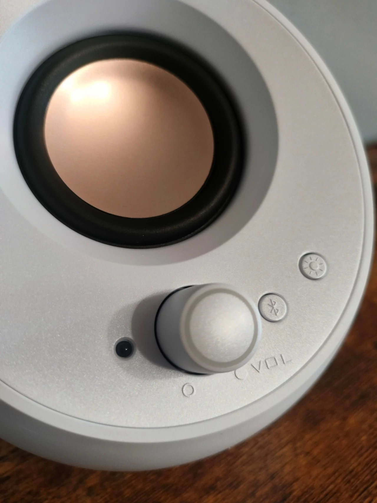 Biały, nowoczesny głośnik z okrągłym przyciskiem regulacji głośności, przyciskiem Bluetooth oraz wskaźnikiem LED, ustawiony na drewnianym blacie.