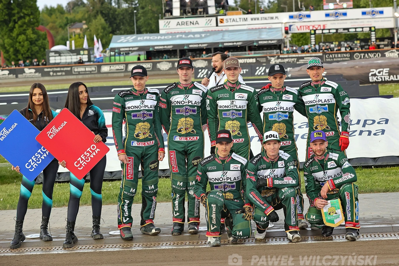Grupa motocyklistów w zielonych kombinezonach z logo sponsorów stoi w rzędzie na torze żużlowym, obok nich dwie kobiety trzymające tablice z nazwami miast, w tle widoczna trybuna i banery reklamowe.