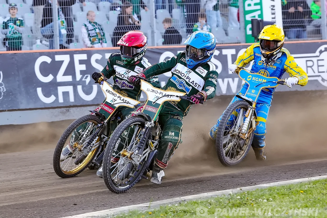 Wiktor Lampart (kask niebieski), Jason Doyle (kask czerwony), Anders Thomsen (kask żółty) Trzech żużlowców w kolorowych kombinezonach i kaskach ściga się na torze, wzbijając kurz podczas dynamicznego zakrętu, na trybunach widoczna jest grupa kibiców za metalową siatką.