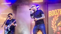 Dwóch muzyków występujących na scenie podczas koncertu, jeden gra na gitarze, drugi śpiewa do mikrofonu, w tle duże podświetlone logo zespołu oraz światła sceniczne.