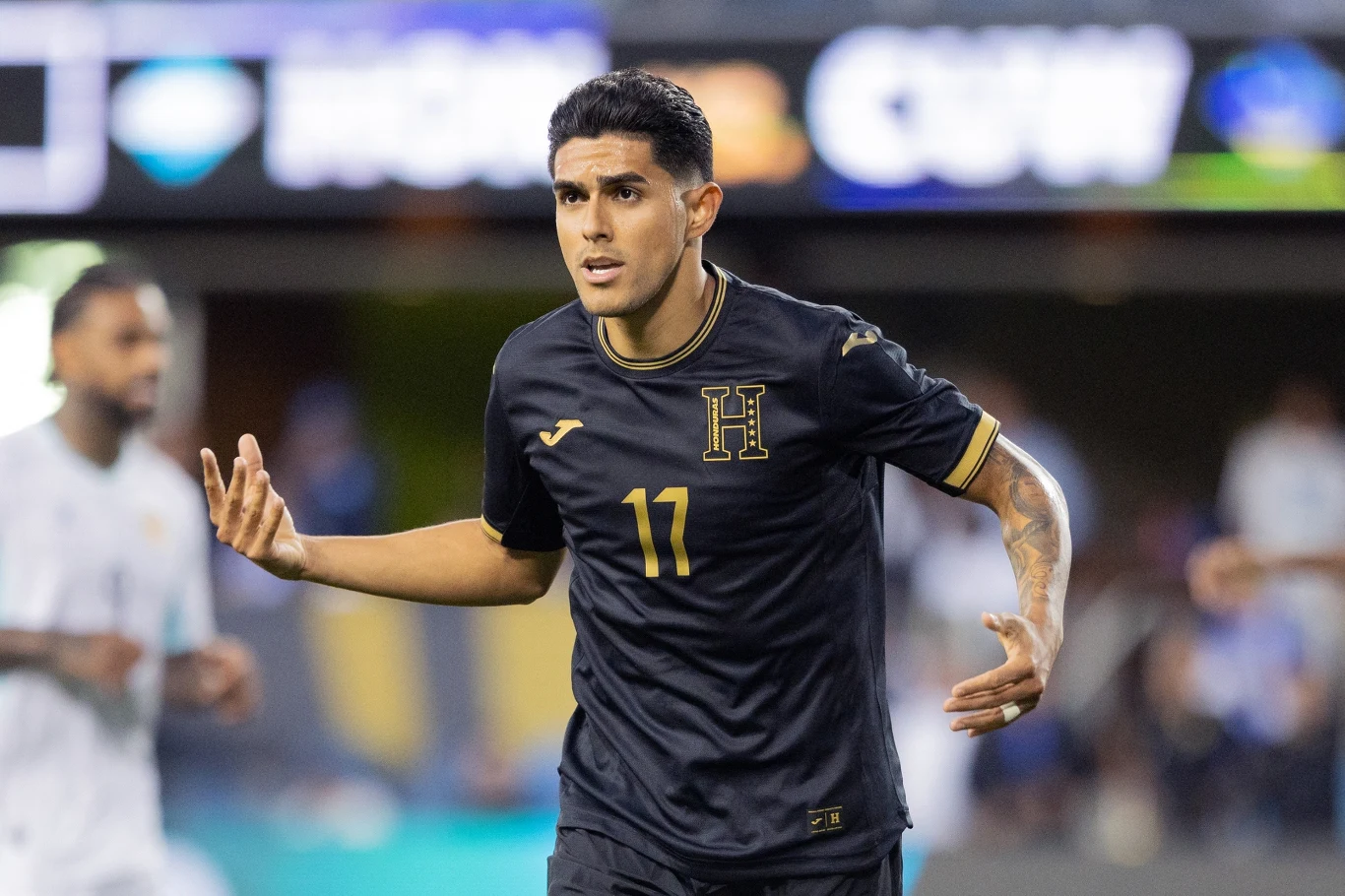 Piłkarz w czarnym stroju z numerem 17 oraz literą 'H' na koszulce celebruje akcję podczas meczu piłkarskiego, za nim rozmazane sylwetki innych zawodników i stadionowe światła.