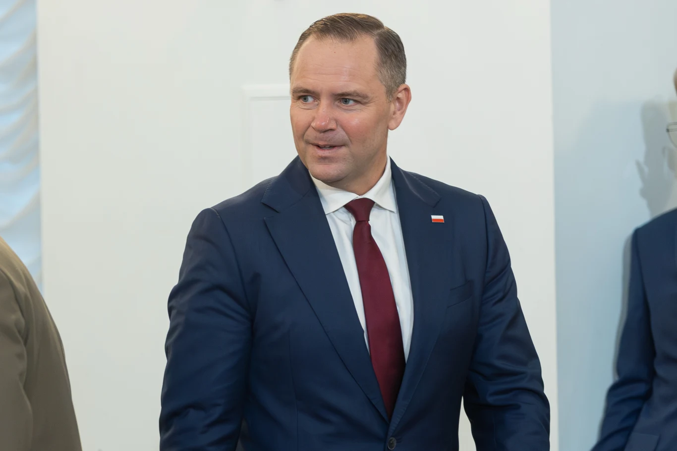 Prezydent elekt Karol Nawrocki Mężczyzna w granatowym garniturze i bordowym krawacie z przypinką w barwach polskiej flagi, uśmiechnięty, w formalnym otoczeniu, skierowany w prawo.