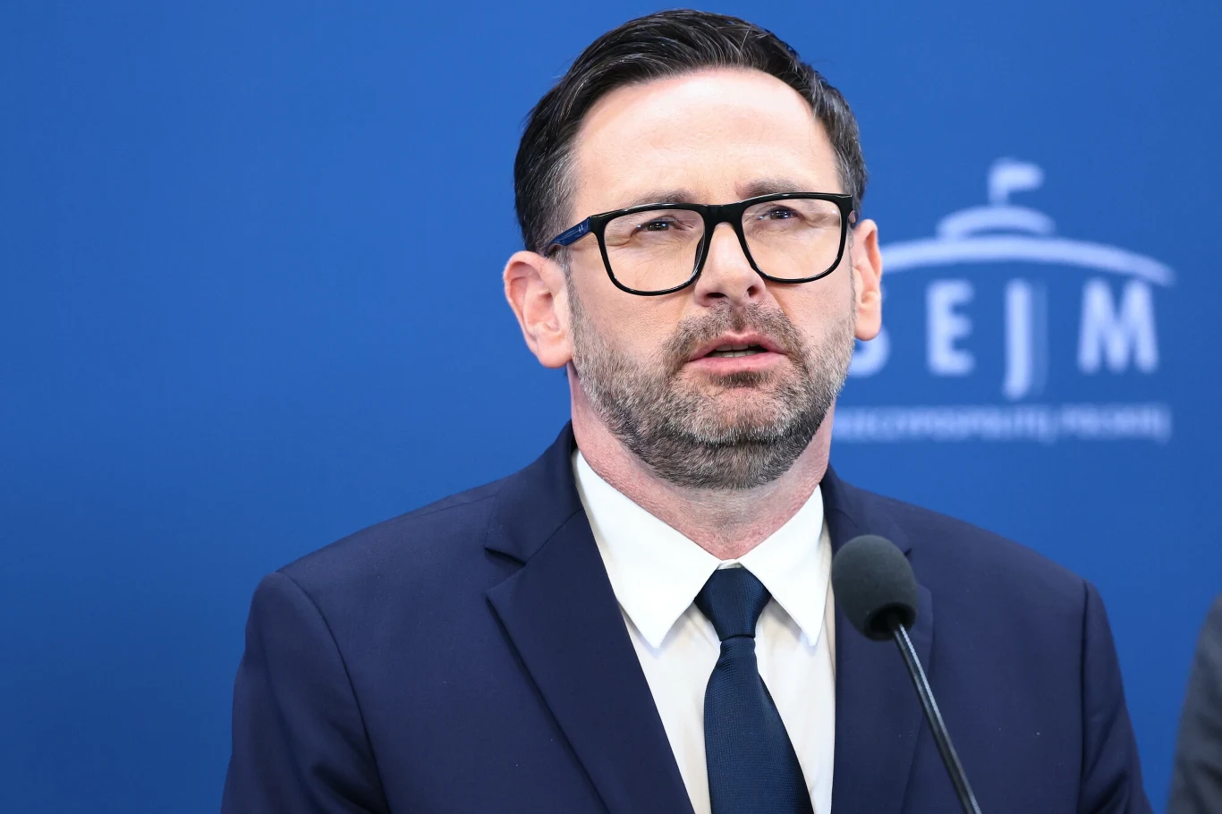 Nowe informacje ws. Daniela Obajtka. Adam Bodnar podpisał wniosek do szefowej PE Mężczyzna w garniturze i okularach wygłaszający przemówienie przed mikrofonem na tle niebieskiej ścianki z napisem SEJM.