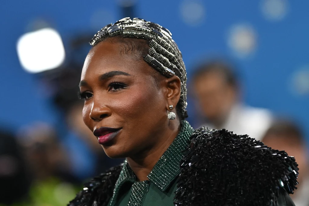 Venus Williams Stylowo ubrana kobieta z krótkimi włosami ozdobionymi metalicznymi klamrami, wyrazistym makijażem i kolczykami, ujęta na tle rozmytego niebieskiego tła z delikatnie widocznymi światłami.