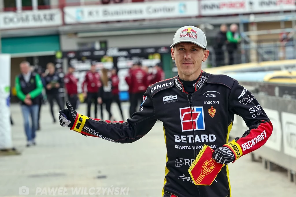 Maciej Janowski Zawodnik żużlowy w stroju drużynowym z licznymi logotypami sponsorów trzyma kask w ręce i pokazuje gest kciuka do góry na stadionie żużlowym, w tle widocznych jest kilka osób związanych z obsługą imprezy.