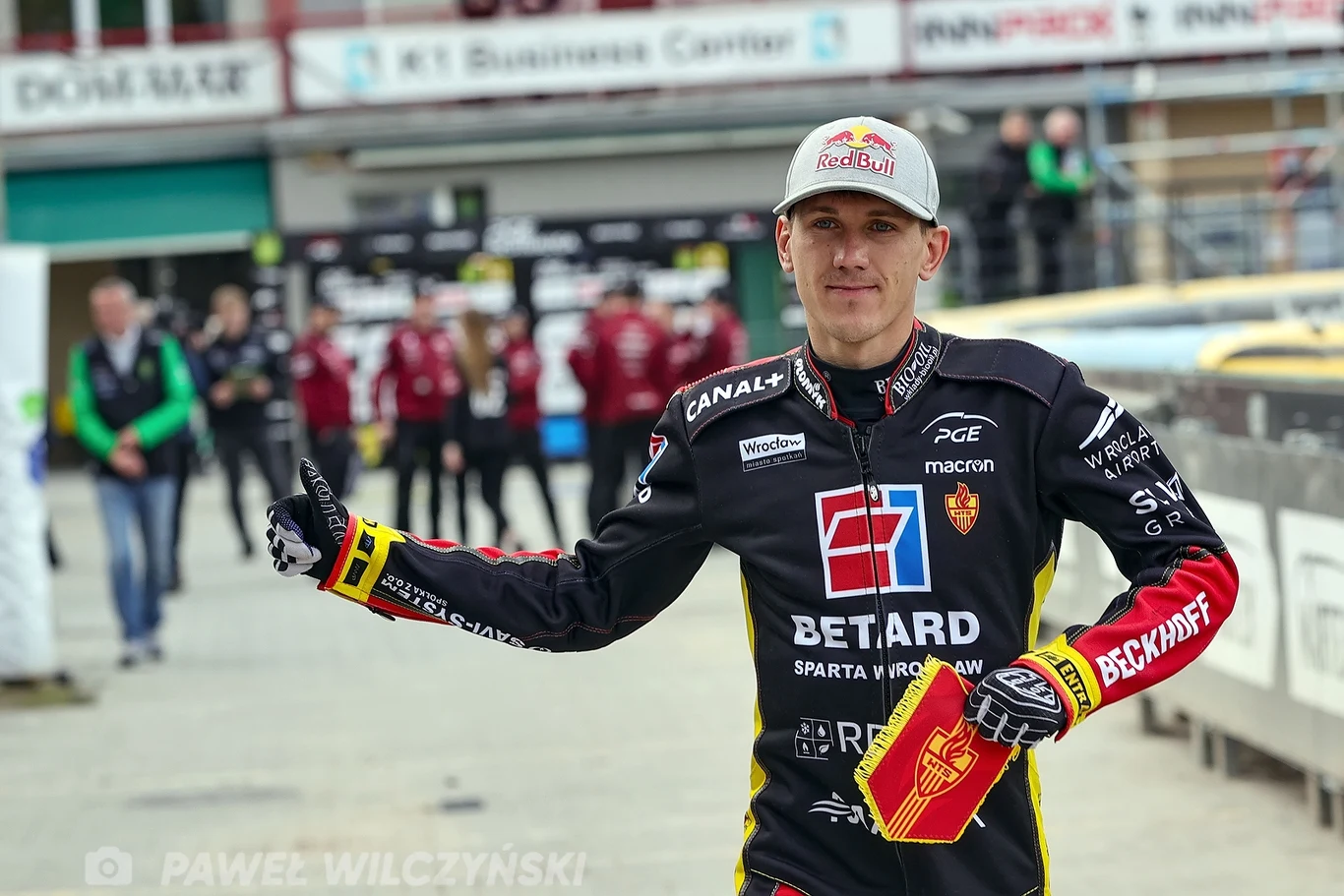 Maciej Janowski Zawodnik żużlowy w stroju drużynowym z licznymi logotypami sponsorów trzyma kask w ręce i pokazuje gest kciuka do góry na stadionie żużlowym, w tle widocznych jest kilka osób związanych z obsługą imprezy.