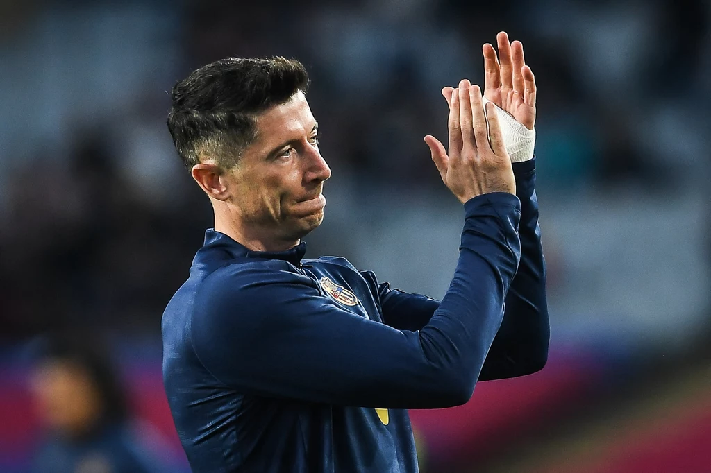 Robert Lewandowski Piłkarz w granatowej bluzie sportowej klaszcze w dłonie, wyrażając podziękowanie lub uznanie, jego dłoń owinięta jest bandażem, tło rozmazane, skupienie na sylwetce sportowca.