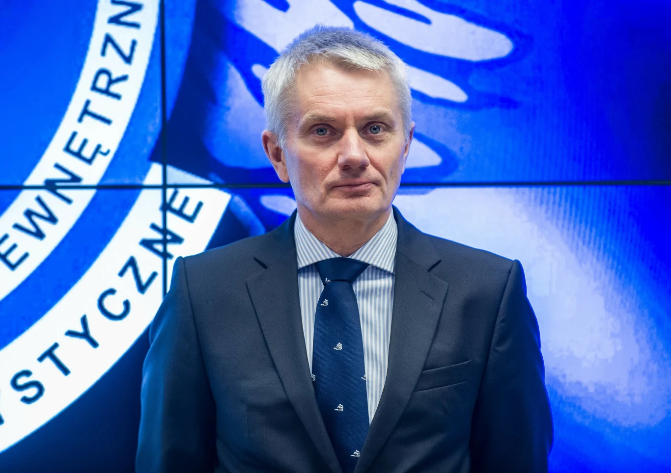 Maciej Hunia nowym ambasadorem RP w Izraelu Mężczyzna w garniturze i krawacie stoi na tle niebieskiego ekranu z fragmentem logo instytucji lub organizacji zawierającego biały napis wokół okręgu.