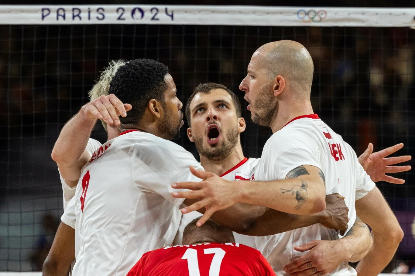 Grupa siatkarzy w białych koszulkach świętuje zdobycie punktu podczas meczu kwalifikacyjnego do Igrzysk Olimpijskich w Paryżu 2024, widoczna siatka oraz napis na górze z nazwą olimpiady.