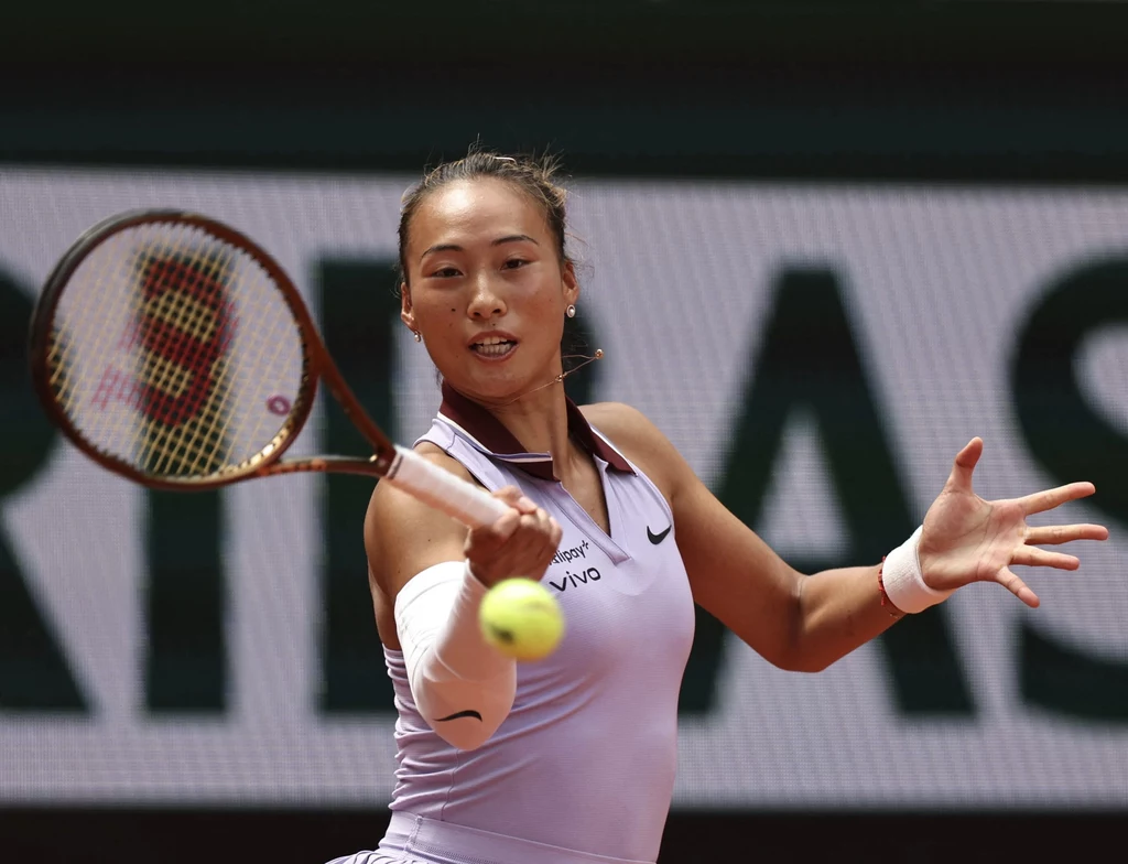 Qinwen Zheng Tenisistka wykonująca zamach rakietą podczas dynamicznej wymiany, skupiona na lecącej piłce tenisowej, w tle rozmazany napis reklamowy.