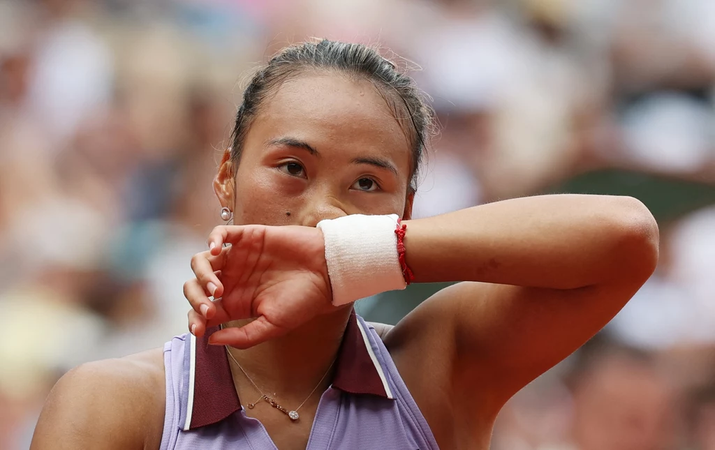 Qinwen Zheng Tenisistka w sportowej odzieży z opaską na nadgarstku ociera czoło przedramieniem, na jej twarzy widać zmęczenie, tło zamazane sugeruje obecność widowni na korcie.