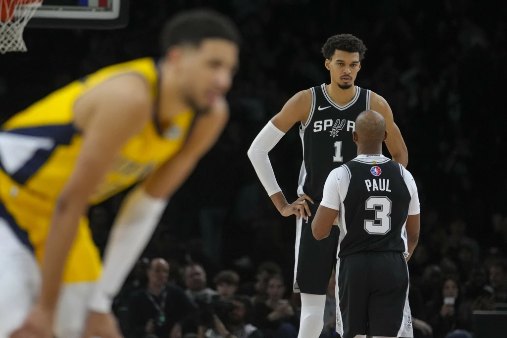 Chris Paul i Victor Wembanyama Dwóch koszykarzy z drużyny San Antonio Spurs i Golden State Warriors stoi naprzeciw siebie w centrum boiska, jeden z nich znacząco przewyższa wzrostem przeciwnika, w tle widoczni kibice i rozmyty zawodnik w żółtej koszulce.