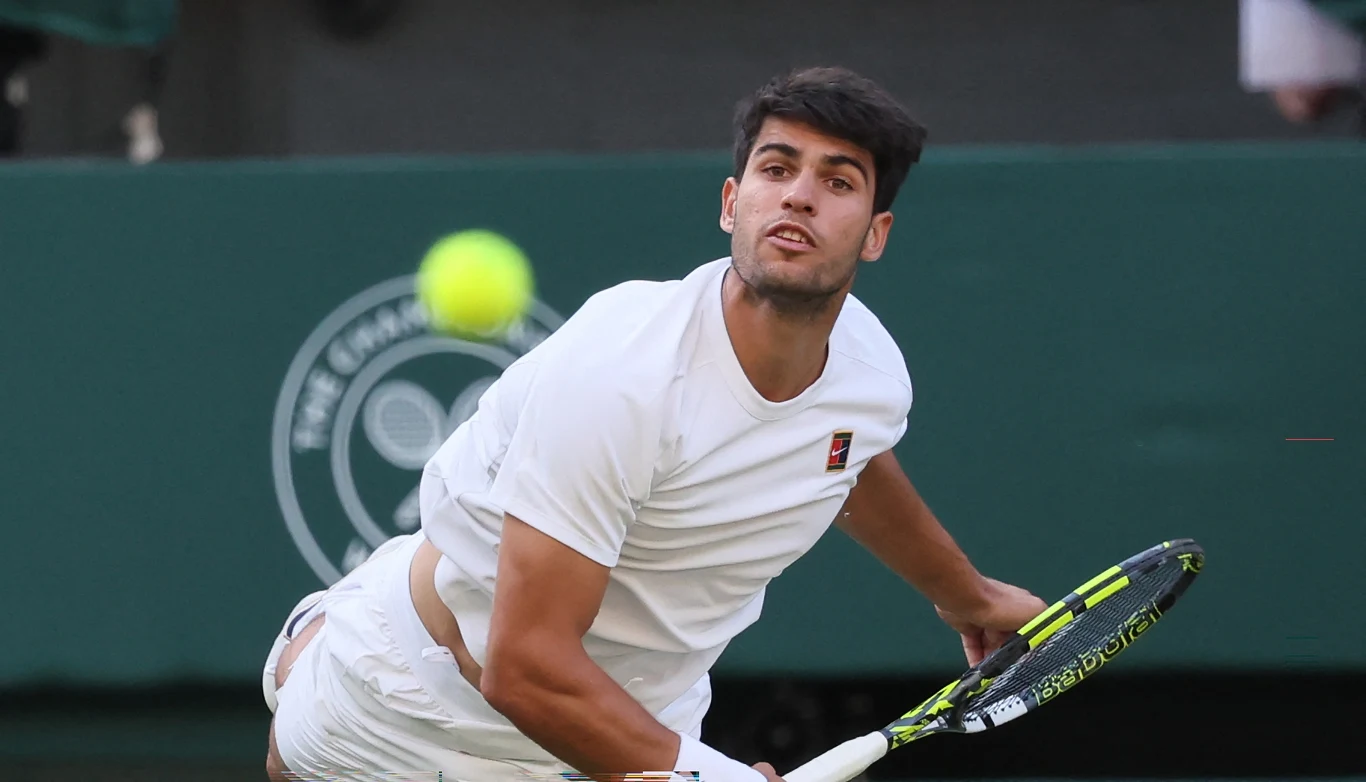 Młody tenisista w białym stroju podczas dynamicznego serwisu na tle zielonego kortu tenisowego z widocznym logo turnieju.