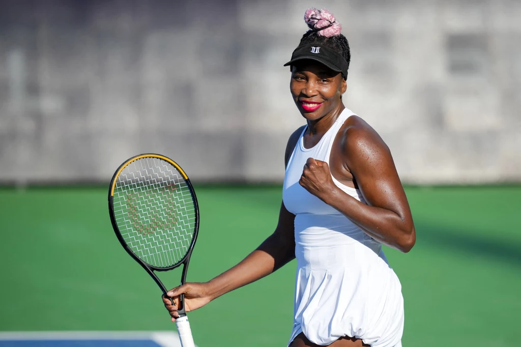 Venus Williams wróciła do zawodowej rywalizacji po kilkunastu miesiącach przerwy Kobieta w białym stroju tenisowym z rakietą w dłoni, uśmiecha się i zaciska pięść w geście triumfu na korcie tenisowym.