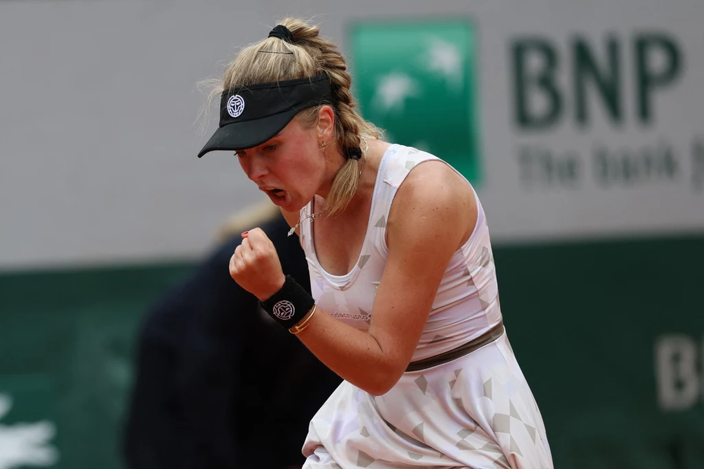 Magdalena Fręch Zawodniczka tenisowa z zaciśniętą pięścią wyraża radość i determinację podczas meczu, ubrana w biały strój sportowy i czapkę z daszkiem, w tle widoczne rozmazane logo sponsora.