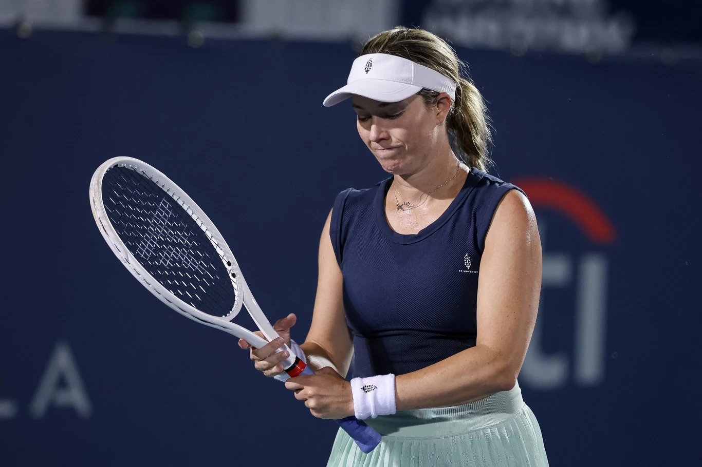 Kobieta w sportowej odzieży i białym daszku trzyma rakietę tenisową, wygląda na skoncentrowaną lub zawiedzioną, tło rozmyte z elementami kortu tenisowego.