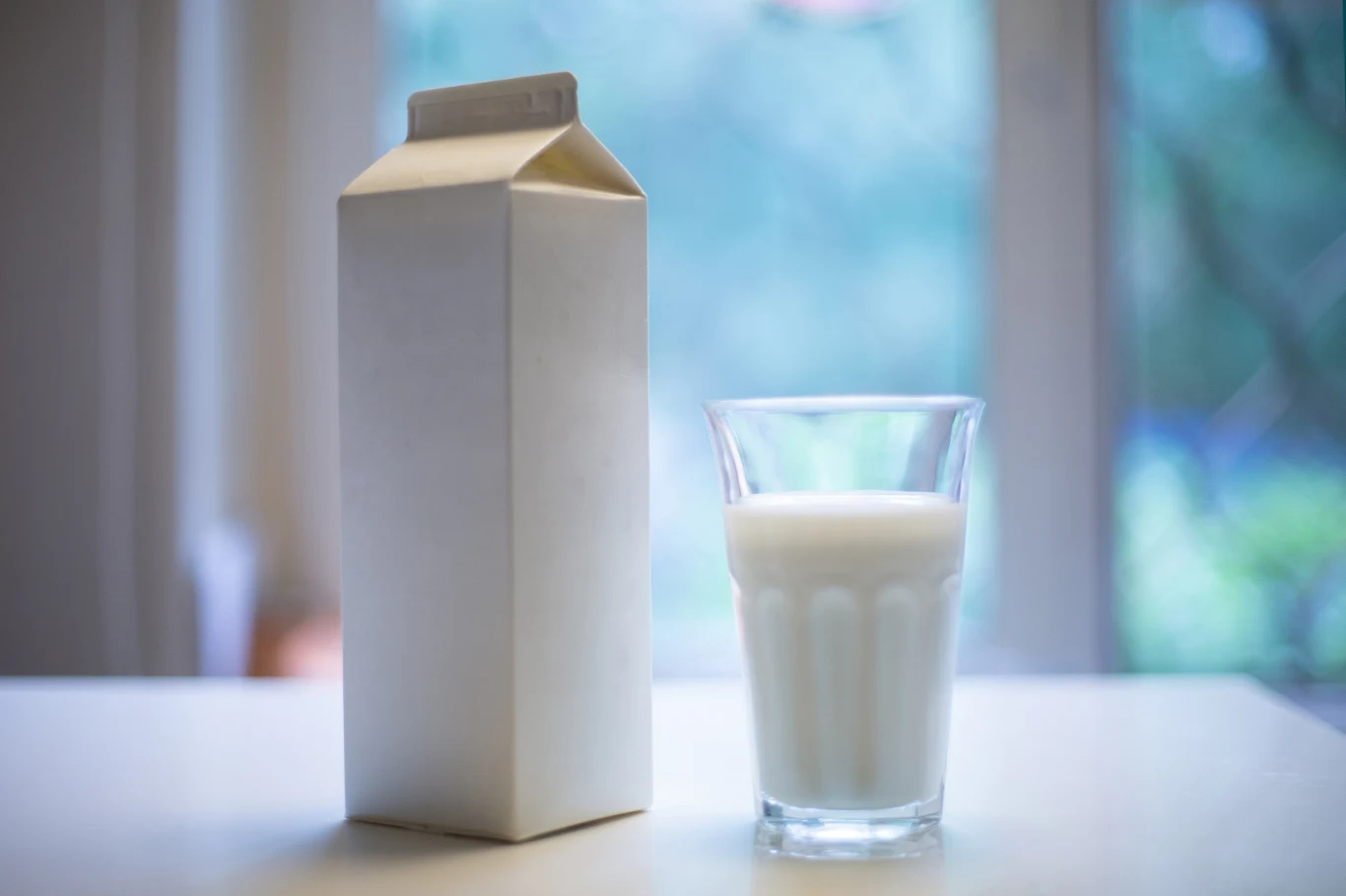 Karton po mleku wyrzucasz do kontenera na papier? Popełniasz ogromny błąd Szklanka napełniona mlekiem ustawiona obok białego, kartonowego opakowania na mleko na jaśniej powierzchni, w tle rozmyte okno z zielonym widokiem.