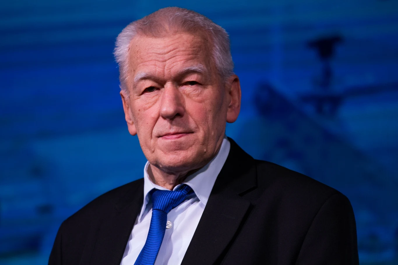 Kornel Morawiecki założył Wolnych i Solidarnych w 2016 r. Starszy mężczyzna w eleganckim garniturze i niebieskim krawacie, na tle rozmazanego niebieskiego tła, patrzący spokojnym wzrokiem w stronę aparatu.