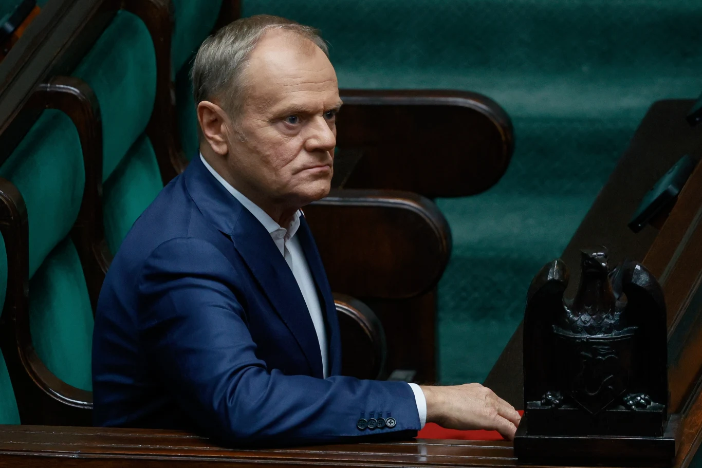 Niepokojący sondaż dla rządu Donalda Tuska Mężczyzna w garniturze siedzi samotnie na ławie w parlamencie, spoglądając poważnie przed siebie, w tle widoczne są zielone obicia ławek oraz fragment stylizowanej mównicy z orłem.