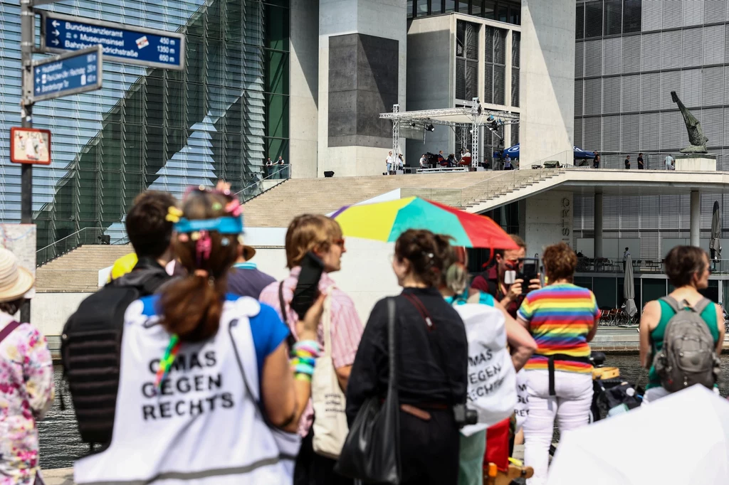 Grupa osób zgromadzonych pod kolorowymi parasolkami, część uczestników ubrana w koszulki z napisem 'OMAS GEGEN RECHTS', w tle nowoczesny budynek i scena z nagłośnieniem.