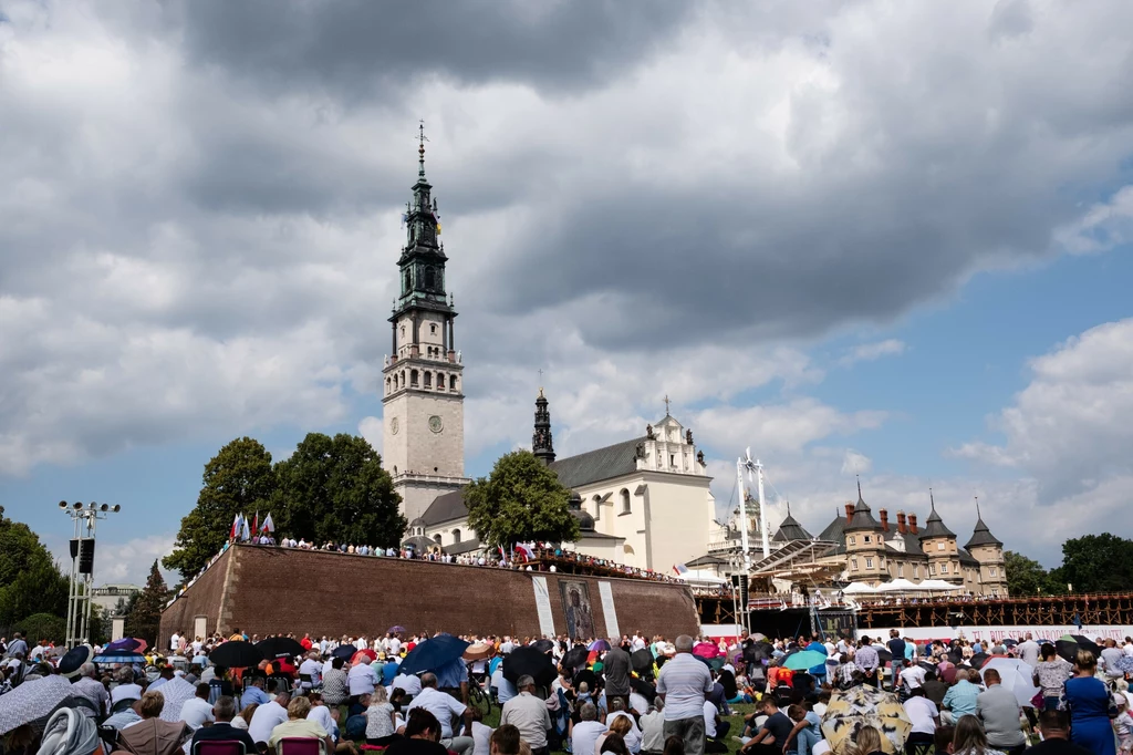 Jasna Góra przyciąga wielu turystów i wiernych Tłum ludzi zgromadzonych przed budynkiem sanktuarium Jasnej Góry w Częstochowie pod pochmurnym niebem, część osób siedzi na trawie, wielu ma ze sobą parasole przeciwsłoneczne, na pierwszym planie podstawowy element stanowi monumentalna bazylika z chara...