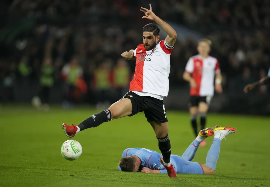 Alireza Jahanbakhsh Dynamiczna akcja podczas meczu piłkarskiego, zawodnik drużyny w biało-czerwono-czarnym stroju wykonuje wysokie podbicie piłki, podczas gdy inny gracz leży na murawie, trzymając się za głowę
