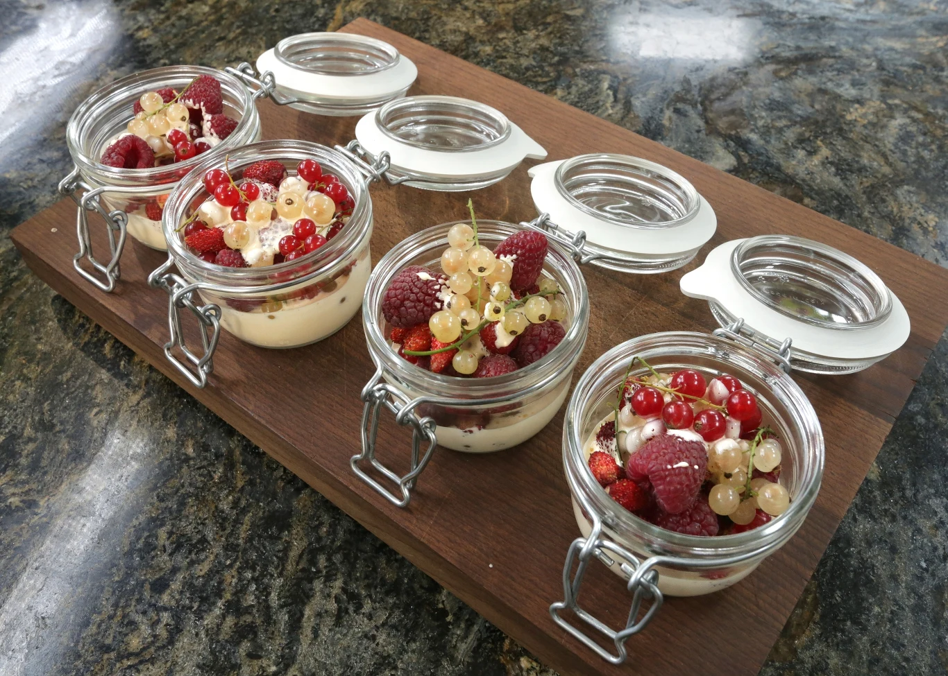 Porzeczki z koglem-moglem Cztery słoiki z zamykanymi wieczkami ustawione na drewnianej desce, wypełnione kremowym deserem, udekorowane malinami, czerwonymi i białymi porzeczkami, całość na kamiennym blacie.