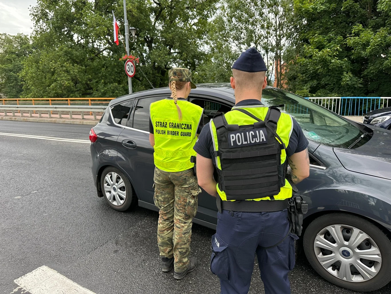 Kontrola na granicy z Niemcami Funkcjonariusze Straży Granicznej oraz Policji przeprowadzają kontrolę samochodu osobowego na ulicy, obok widoczny znak ograniczenia prędkości do 30 km/h oraz barierka i drzewa w tle.