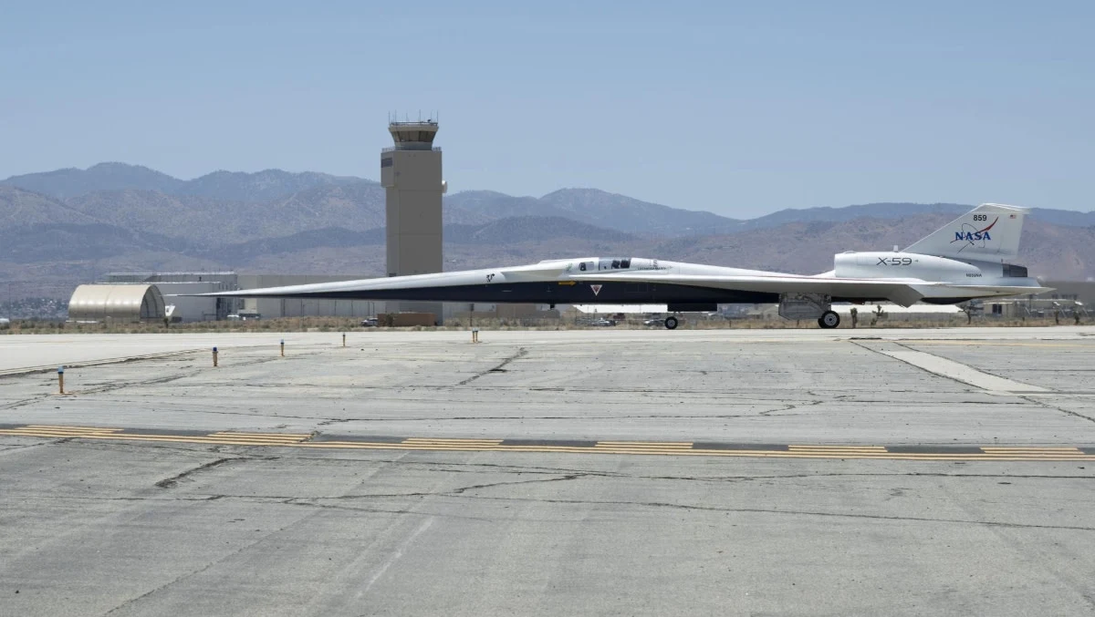 NASA testuje naddźwiękowy odrzutowiec X-59. Kiedy pierwszy lot? Eksperymentalny samolot NASA X-59 stoi na płycie betonowego lotniska, w tle widoczna wieża kontroli lotów oraz masyw górski pod bezchmurnym niebem.