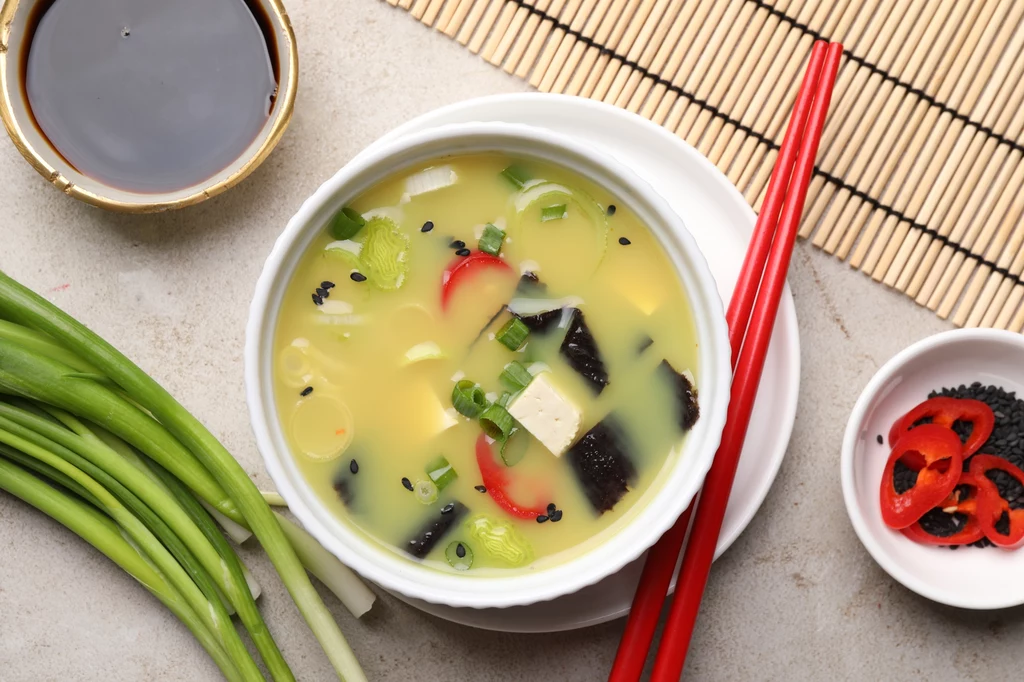 Miska zupy miso z dodatkiem tofu, wodorostów, czarnego sezamu, plasterkami czerwonej papryki oraz posiekanymi szczypiorkami, u boku pałeczki, świeża cebulka dymka oraz miseczka z sosem sojowym i pokrojonymi papryczkami chili.