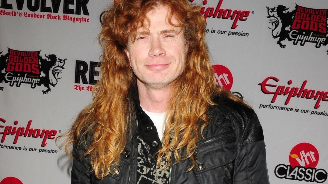 Dave Mustaine (Megadeth) Mężczyzna z długimi rudymi włosami w czarnej kurtce skórzanej stoi na tle ścianki promocyjnej z logotypami marek i wydarzeń muzycznych.
