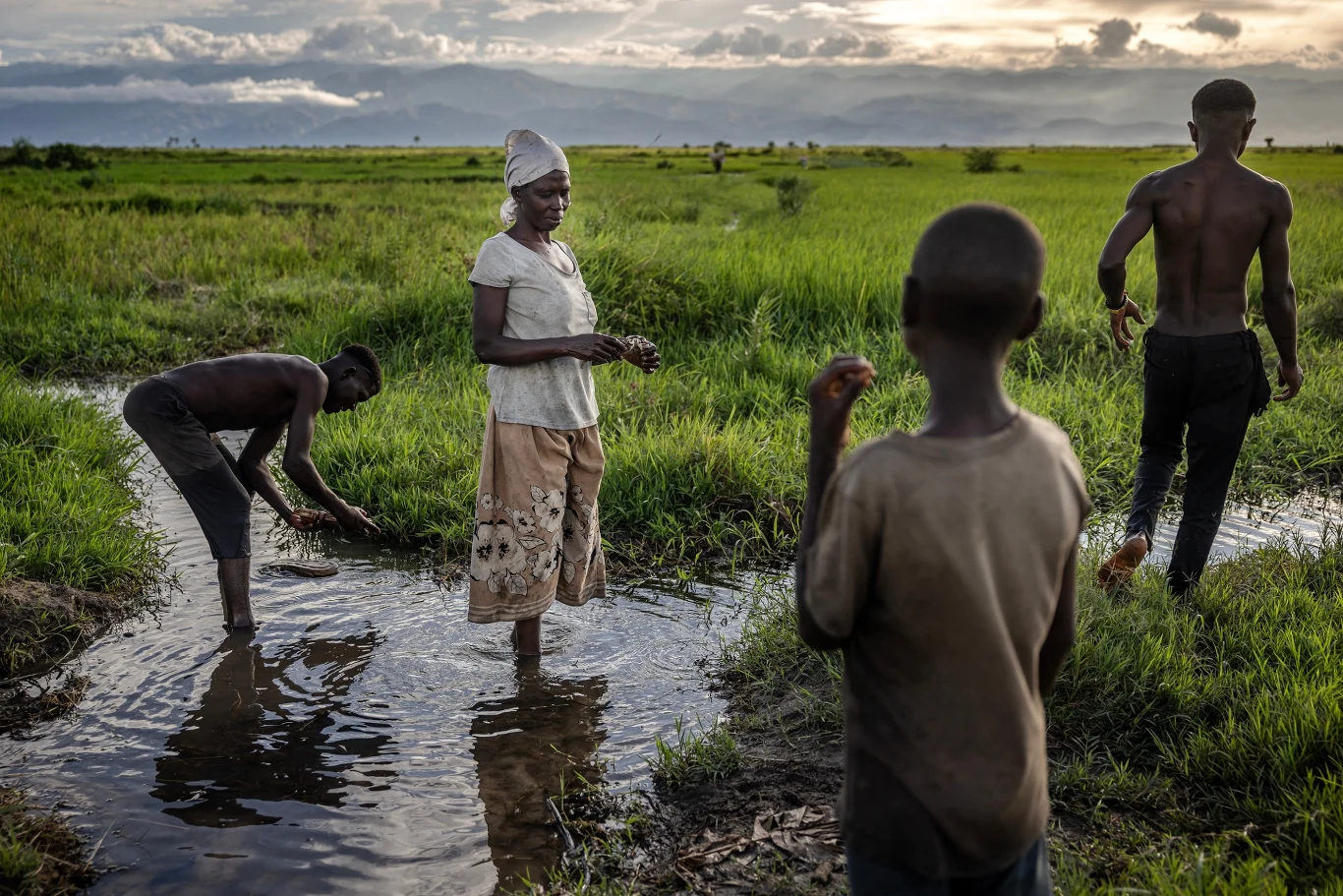 Cztery osoby, w tym kobieta i trzech chłopców, stoją w płytkiej wodzie otoczonej zielonymi polami, w tle rozciągają się góry i zachmurzone niebo, dominuje spokojna, naturalna sceneria.