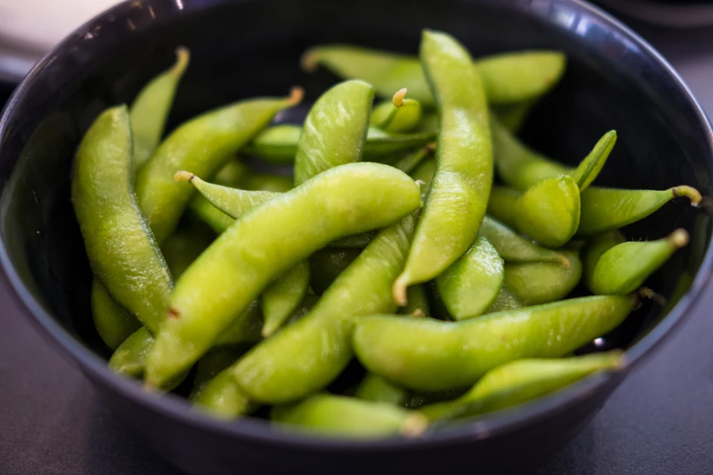 Fasolę edamame można potraktować jako warzywny dodatek do obiadu Świeże, zielone strąki edamame w czarnej misce, kropelki wody widoczne na powierzchni fasolek, kontrastujące z ciemnym tłem.