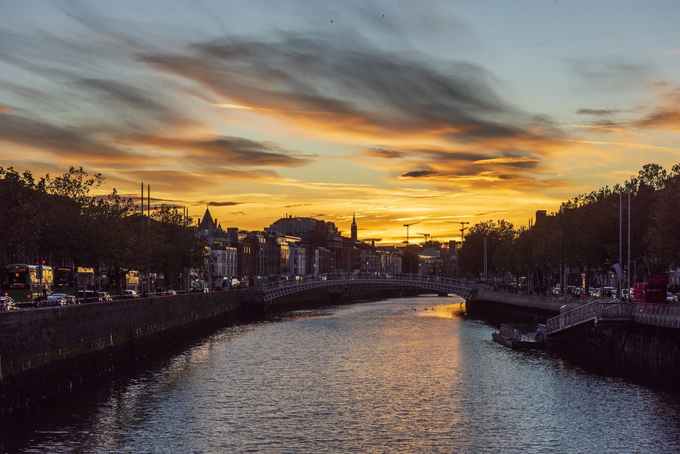 Most rozciąga się nad rzeką w centrum miasta, a po obu stronach widoczne są budynki oraz drzewa. Nad horyzontem rozciąga się spektakularny zachód słońca, którego barwy odbijają się na powierzchni wody.