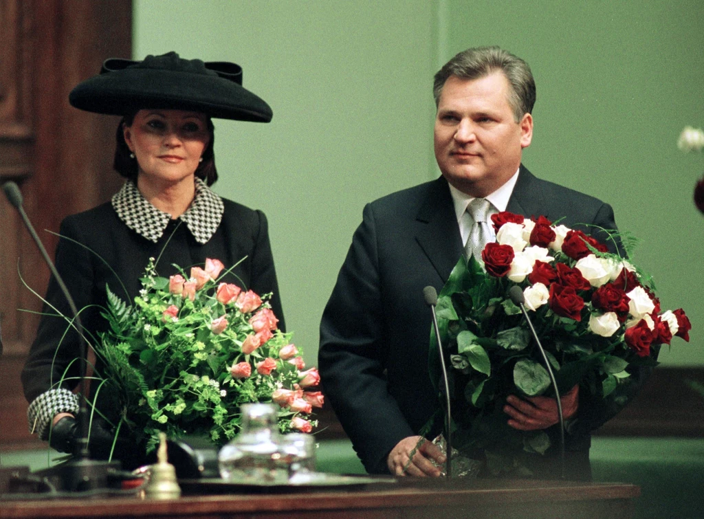 Para dorosłych w eleganckich, formalnych strojach stoi w budynku, każde z nich trzyma duży bukiet kwiatów, kobieta ubrana w czarny kapelusz i kostium z kołnierzem w kratę, mężczyzna w ciemnym garniturze.