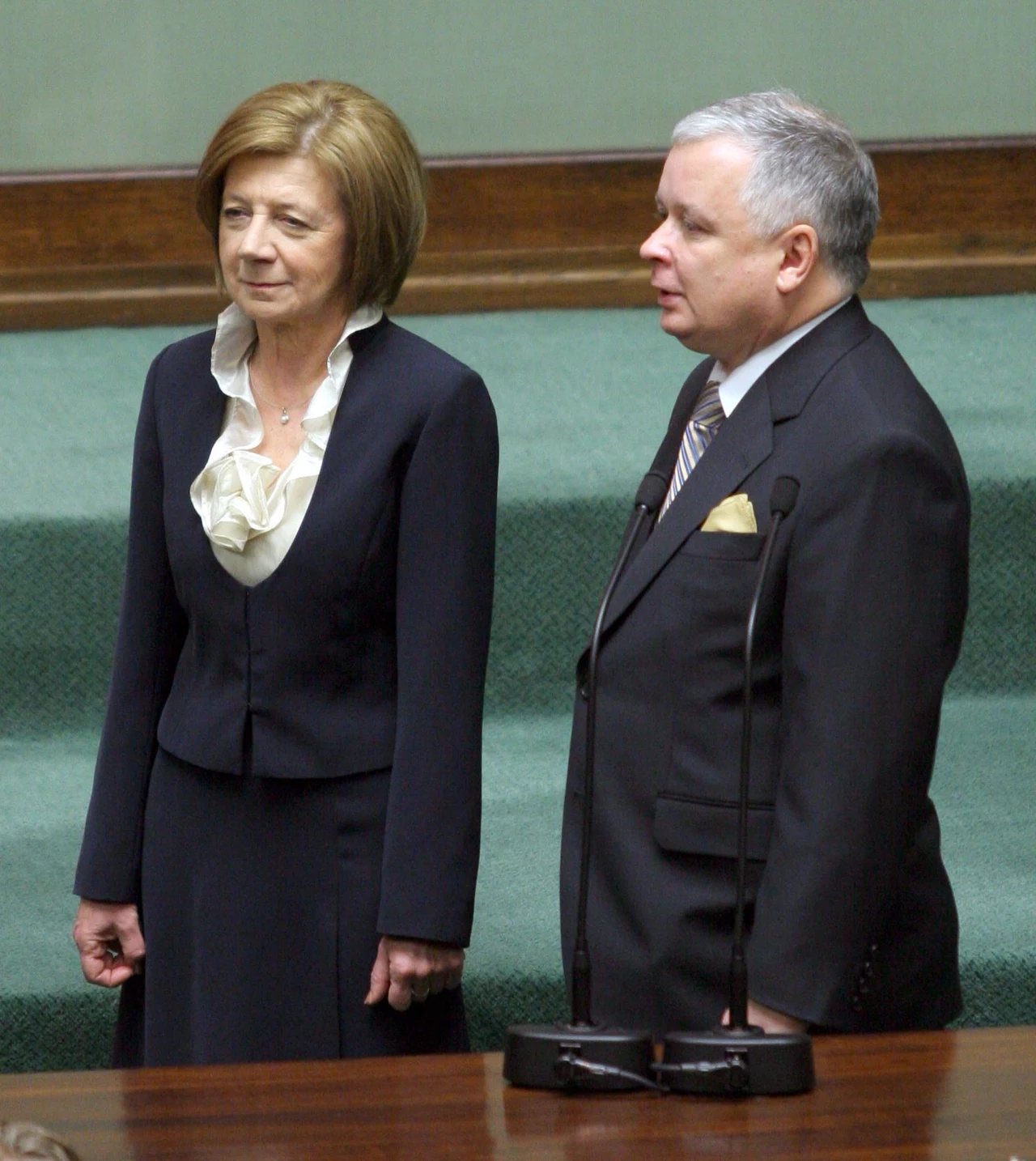 Dwie osoby stojące obok siebie w eleganckich, formalnych ubraniach na tle zielonej sali z mównicą, po lewej kobieta w garniturze z jasną bluzką, po prawej mężczyzna w garniturze z poszetką, oboje zwróceni w stronę mównicy.