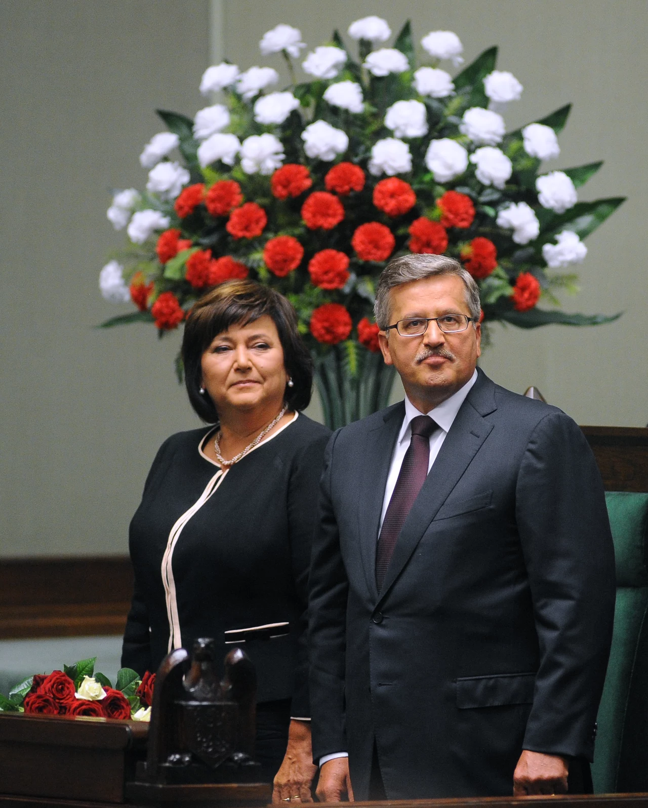 Dwie elegancko ubrane osoby stojące na tle okazałego bukietu kwiatów w barwach czerwono-biało-zielonych, znajdujące się w przestrzeni oficjalnej, prawdopodobnie w sali sejmowej, obok bukiet róż.