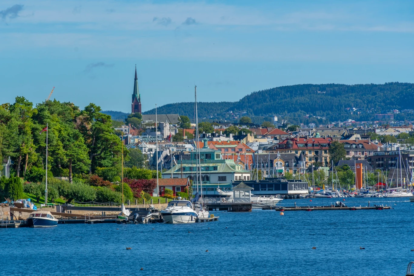 Widok miasta położonego nad wodą z przystanią dla jachtów, otoczony zielenią, w tle rozciągają się wzgórza oraz widoczna jest wysoka wieża kościoła.
