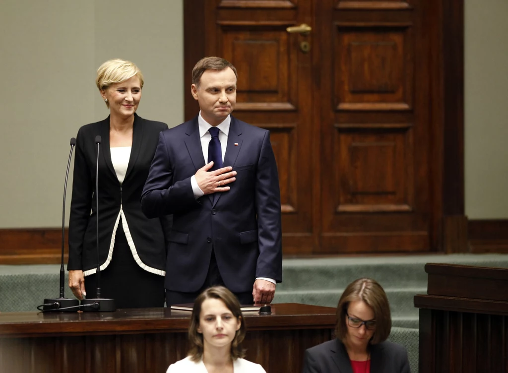 Mężczyzna w garniturze z dłonią na sercu stoi obok kobiety w formalnym stroju, za nimi drewniane drzwi w sali o oficjalnym charakterze, przed nimi dwie kobiety siedzące przy biurkach.