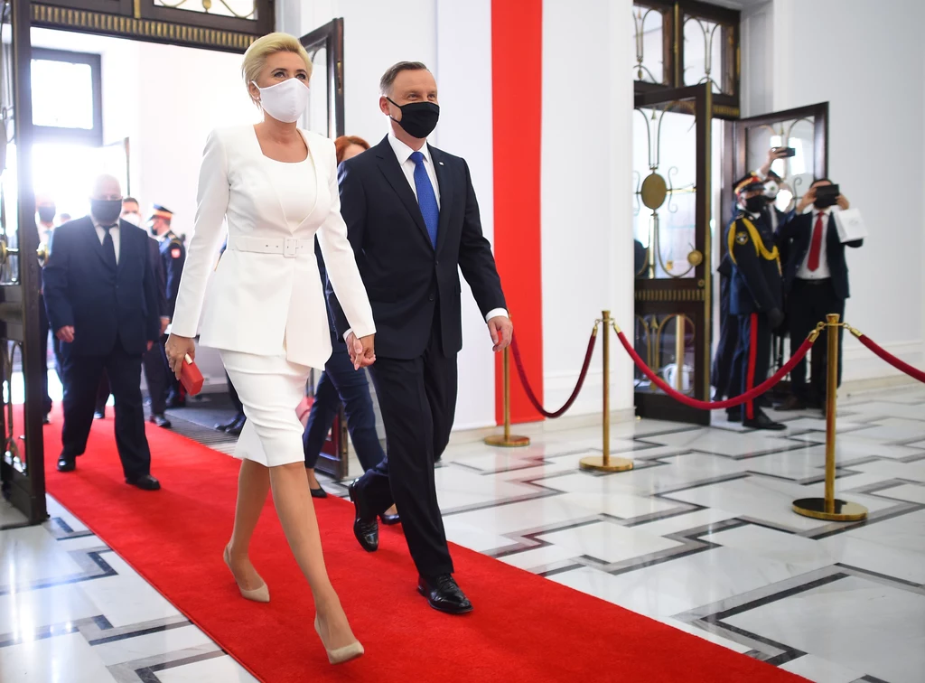 Para ubrana elegancko, w maseczkach, idzie po czerwonym dywanie w przestronnym, oficjalnym wnętrzu z biało-czerwonymi elementami dekoracyjnymi, w tle widoczni funkcjonariusze oraz fotoreporterzy.