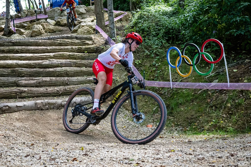 Paula Gorycka Kolarz w białym stroju z czerwonym kaskiem podczas wyścigu górskiego na rowerze, na tle kolorowych pierścieni olimpijskich i leśnego terenu z przeszkodami z drewnianych bali.