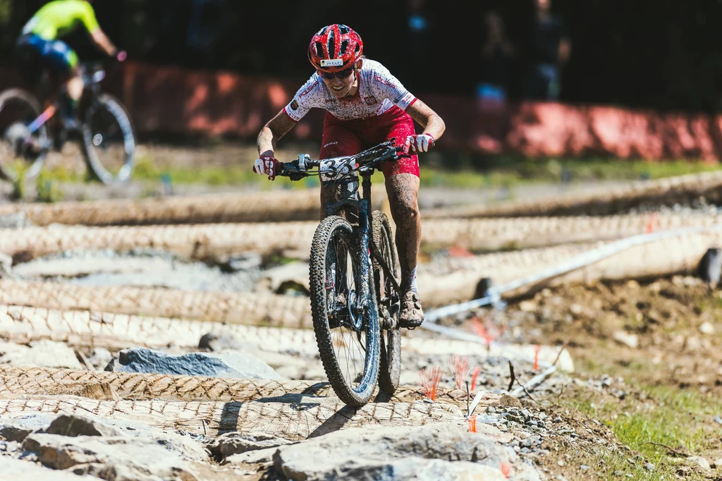 Paula Gorycka Kolarz górski w sportowym stroju jedzie po kamienistej trasie, intensywnie skupiony, otoczony naturalnym terenem, w tle inny zawodnik oraz elementy toru wyścigowego.